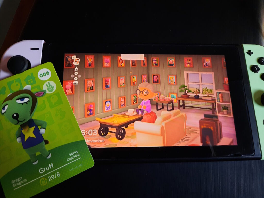 Der angeschaltete Bildschirm einer Switch, der einen Raum in meinem Haus bei Animal Crossing zeigt. Meine Spielfigur sitzt dabei auf einem Pouf an einem Tisch mit einer Tasse Kaffee vor sich. Im Hintergrund sind sehr, sehr viele Fotos von unterschiedlichen Nachbarn an den Wänden zu sehen. Auf dem Bildschirm liegt eine Amiibo-Karte, die die Figur Gregor (ein grüner Ziegenbock) zeigt.