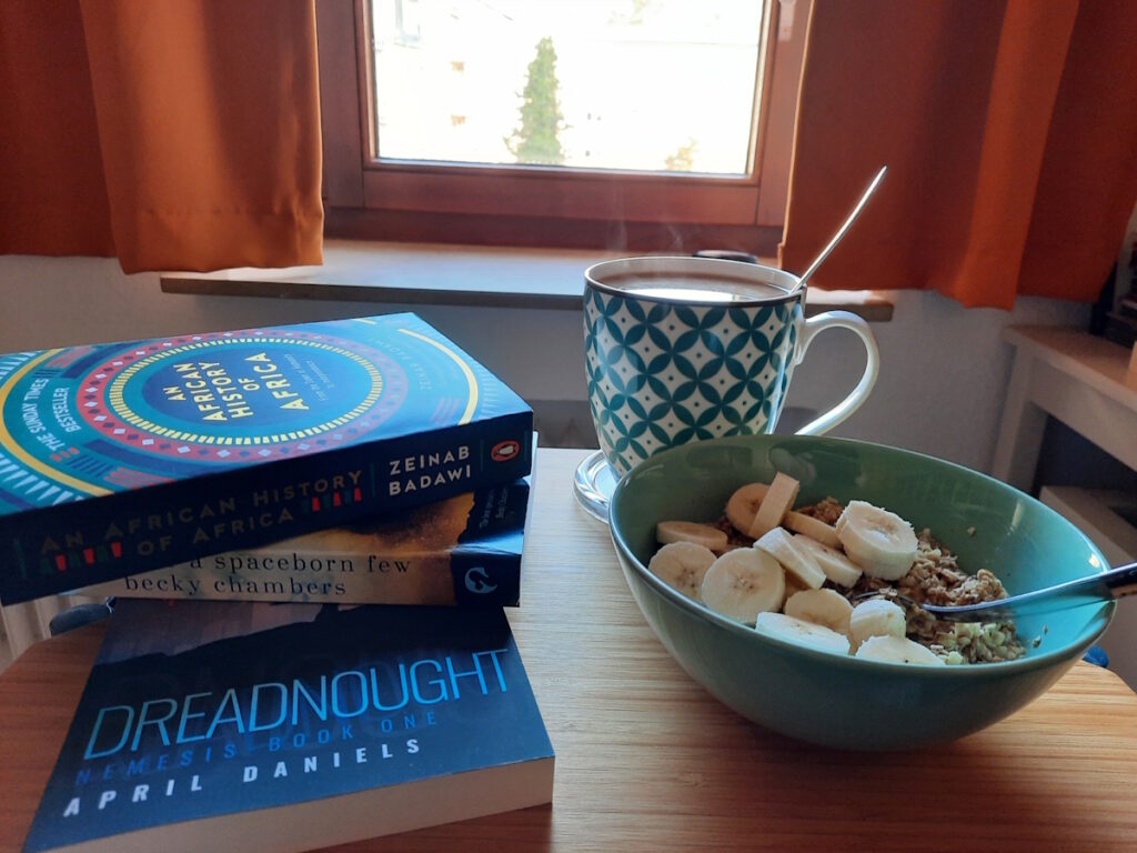 Ein kleiner Tisch mit einer zartgrünen Schale mit Porridge und Bananenscheiben. Dahinter eine grün-weiß-gemusterte Tasse mit dampfendem Tee und links von der Schale ein Stapel Bücher. Die Buchtitel lauten von unten nach oben Dreadnought, Record of Spaceborn Few und An African History of Africa