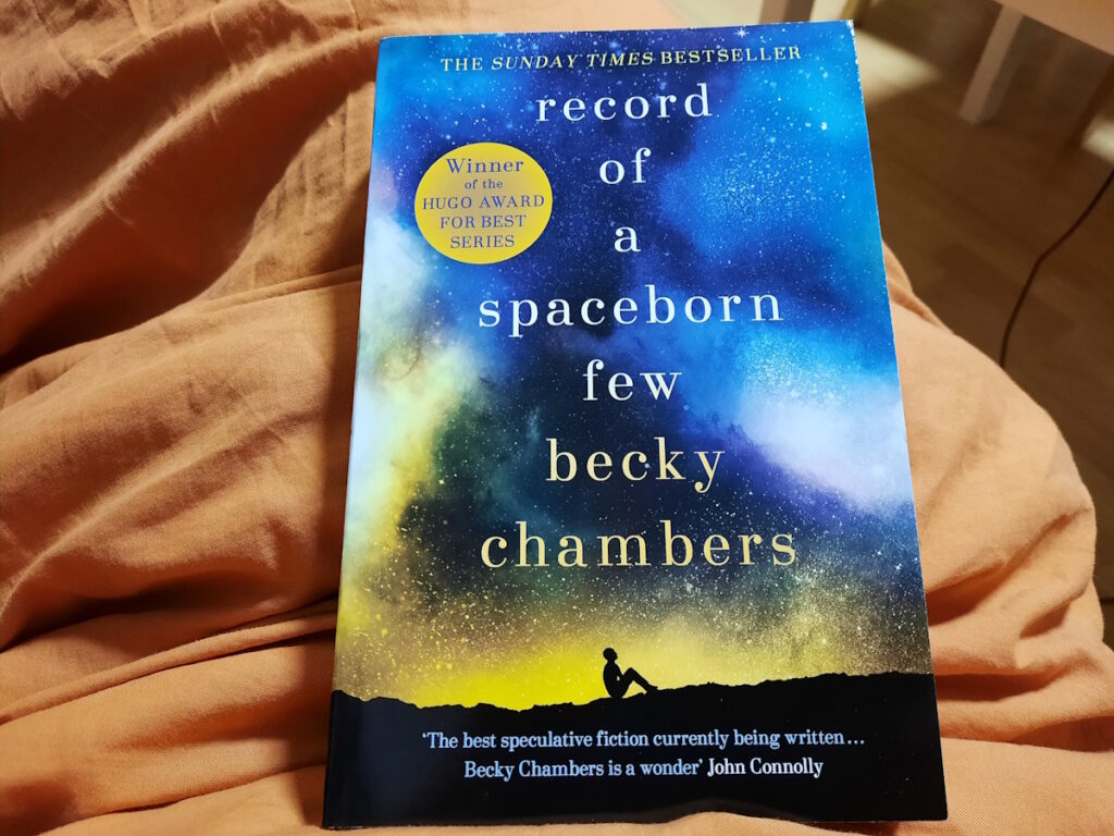 Die Taschenbuchausgabe von "Record of a Spaceborn Few" von Becky Chambers auf einer orangen Decke. Das Cover zeigt einen von blau (oben) nach gelb (unten) verlaufenden Himmel über den in Kleinbuchstaben Titel und Autorenname geschrieben wurden. Ganz unten lässt sich die Silhoutte einer Person erahnen, die auf dem Boden sitzt und in den Himmel schaut.