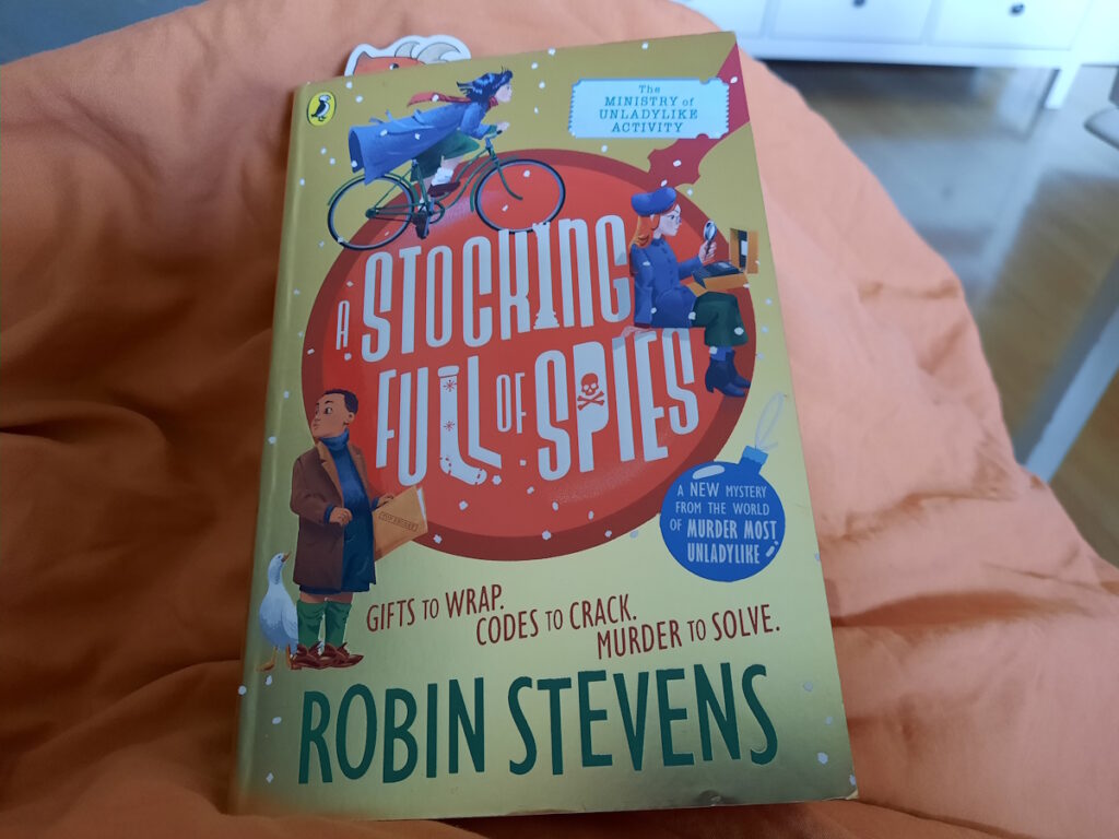 Die Taschenbuchausgabe von "A Stocking Full of Spies" auf einer orangen Decke. Das Cover ist goldfarbend mit einem großen roten Kreis, der an eine Weihnachtsbaumkugel erinnert. In dem Kreis steht der Titel und drumherum sind drei Figuren zu sehen. Oberhalb der Kugel ein dunkelhaariges Mädchen auf einem Fahrrad, rechts von der Kugel ein rothariges Mädchen mit einer Dechiffrier-Maschine und einer Lupe. und unten links ein Junge mit einer Top-Secret-Akte in der Hand und neben ihm steht eine weiße Ente.
