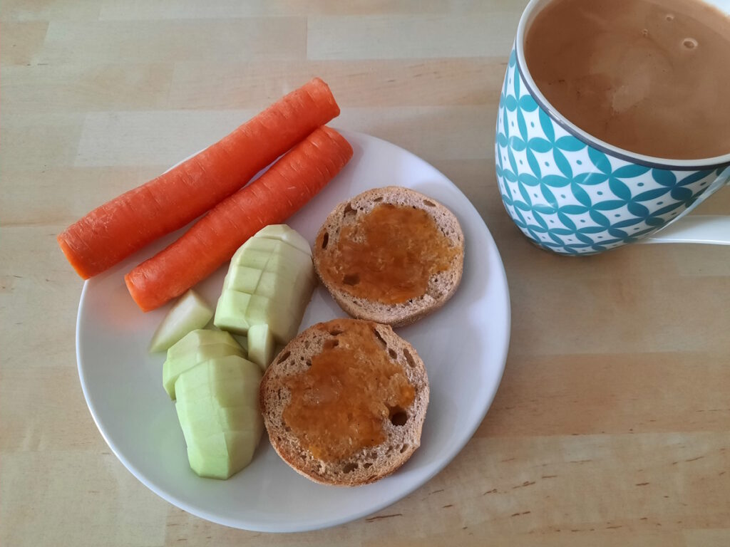 Ein Teller mit einem getoasteten English Muffin aus Sauerteig, der mit Mango-Chutney bestrichen wurde, und zwei Möhren und eine aufgeschnittene kleine Kohlrabi. Daneben eine große Tasse mit Tee.