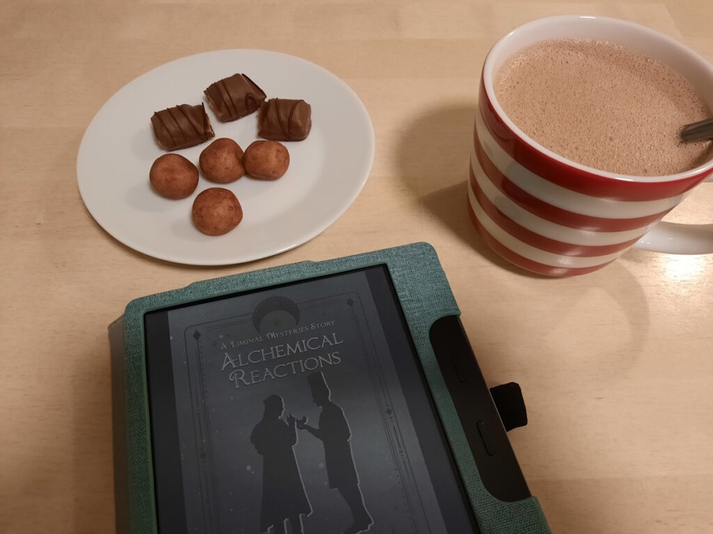 Ein angeschalteter eReader mit dem Cover von "Alchemical Reactions" von Celia Lake. Das Bild zeigt den Schattenriss zweier Figuren in Herrenkleidung, die zueinander zugewandt stehen. Darüber eine Untertasse mit Marzipankartoffeln und Schokostückchen, daneben eine rotweiß-geringelte Tasse mit Heißer Schokolade.