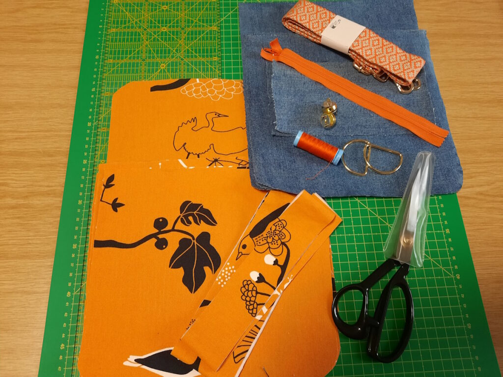 Eine grüne Schneidematte auf der eine Stoffschere, ein oranger Reißverschluss, ein Taschengurt und verschiedene Stoffstücke liegen. Die Stoffstücke sind zum Teil orange mit schwarz-weißem Muster und zum Teil aus Jeansstoff.
