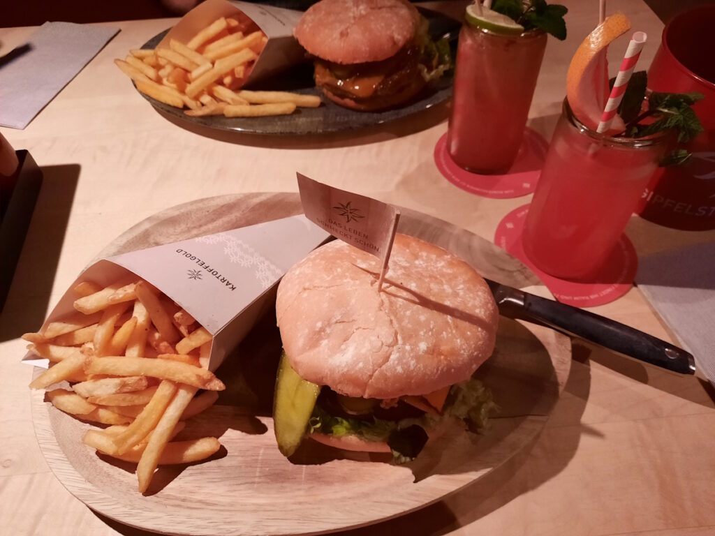 Ein vegetarischer Burger auf einem Holzbrett. Neben dem üppig belegtem Burger liegt eine Spitztüte aus der Pomme Fritz fallen. Neben dem Teller ist eine Grapefruit-Limonade zu sehen.