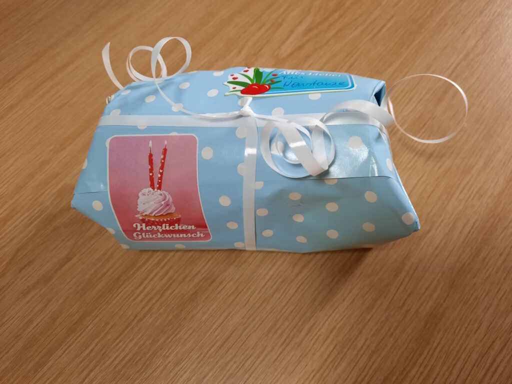 Ein kleines Päckchen in Geschenkpapier. Das Geschenkpapier ist hellblau mit weißen Punkten und darauf klebt ein rosafarbender Aufkleber mit dem Schriftzug "Herzlichen Glückwunsch" und einem Cupcake mit zwei Kerzen drin.