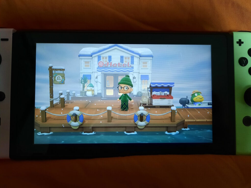 Ein Switch-Bildschirm, auf dem gerade AnimalCrossing läuft. Meine Spielfigur ist in einen grünen, langen Rock gekleidet mit dickem Wollpulli und Mütze und steht auf einem Holzsteg am Meer. Im Hintergrund ist das neue Hotel zu sehen, das es seit dem letzten Update im Spiel gibt.