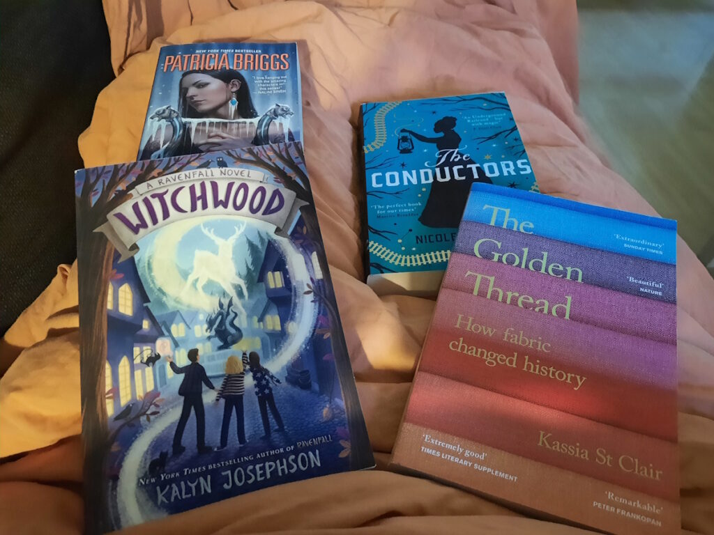 Vier Bücher auf einer orangen Decke: oben links "Winter Lost" von Patricia Briggs, unten links "Witchwood" von Kalyn Josephson, oben rechts "The Conductors" von Nicole Glover, unten rechts "The Golden Thread" von Kassia St. Clair.