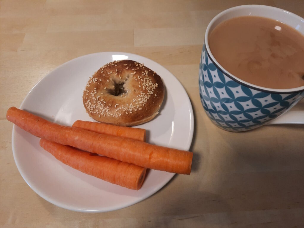Ein weißer Teller mit einem goldbraunem Sesam-Bagel und einigen Möhren. Daneben steht eine große Tasse mit Tee.