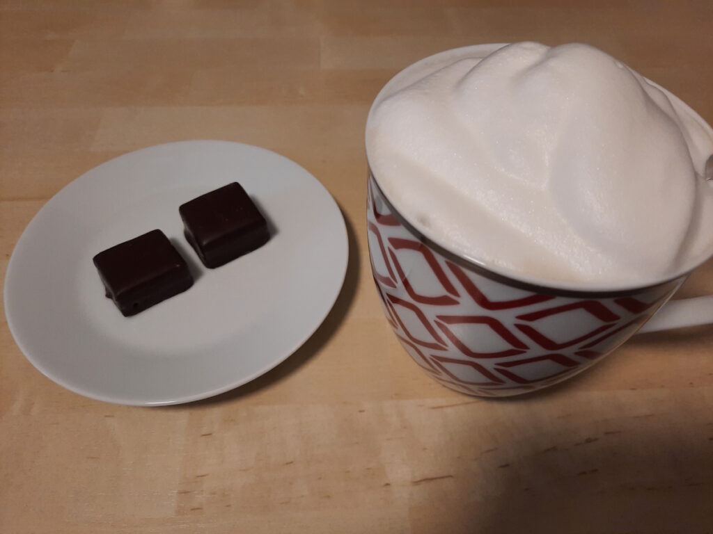 Links eine Untertasse mit zwei schokoladigen Stückchen (genau gesagt Dominosteine) und rechts eine große Tasse mit Milchkaffee.