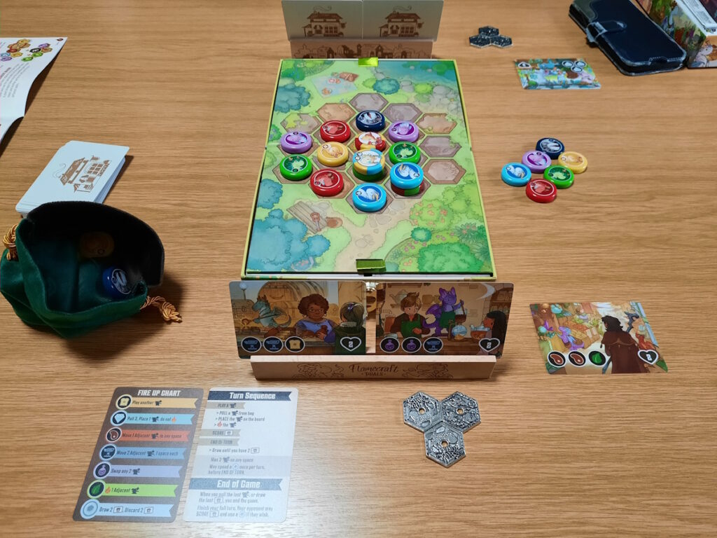 Ein 2-Personen-Spiel von Flamecraft Duals. Im Vordergrund ist eine Halterung mit zwei Karten zu erkennen, sowie drei Metalmünzen und ein Beutel mit Tokens. Hinter den Karten ist ein Spielfeld mit mehreren bunten Tokens zu sehen und dahinter die Rückseite einer weiteren Kartenhalterung.