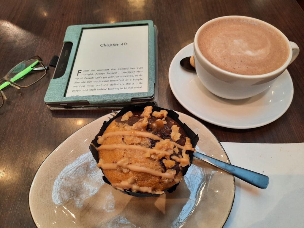Ein goldbrauner Muffin auf einem Teller, daneben eine breite Tasse mit heißem Kakao und ein angeschalteter eReader.