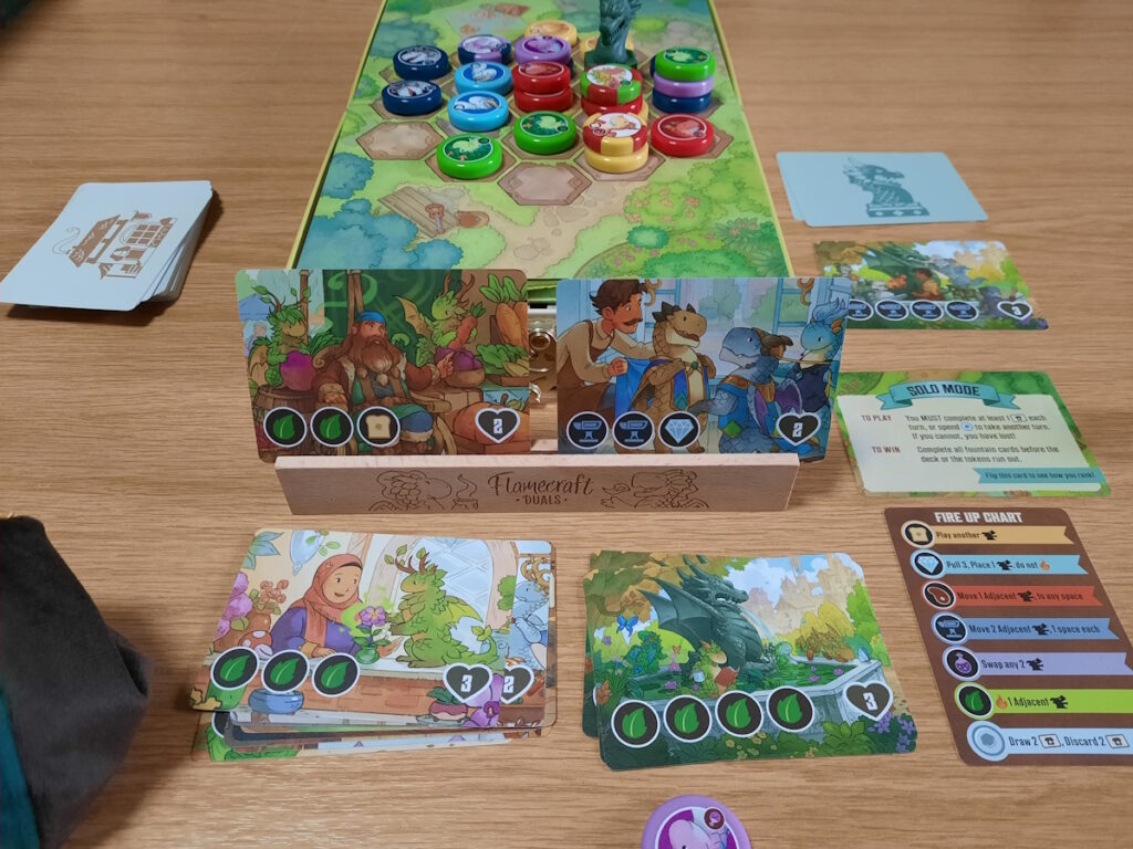 Eine Solo-Partie von Flamecraft Duals, bei der auf dem Spielfeld nicht nur mehrere farbige Tokens, sondern auch ein Plastikbrunnen zu sehen ist. Im Vordergrund steht wieder ein Halter mit zwei Spielkarten, sowie mehrere Stapel mit Karten, die davor lieben.