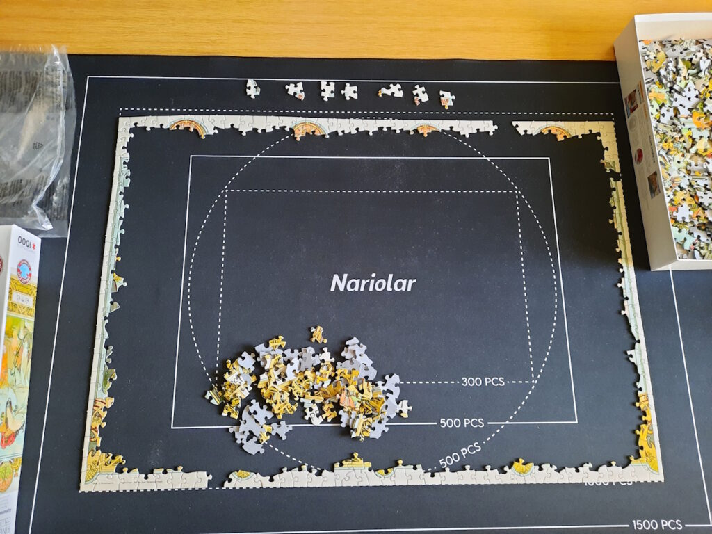 Ein angefangenes Puzzle auf einer Puzzlematte. Bislang ist gerade mal der Rahmen (fast) fertig und eine Handvoll Puzzleteile liegen im Rahmen.