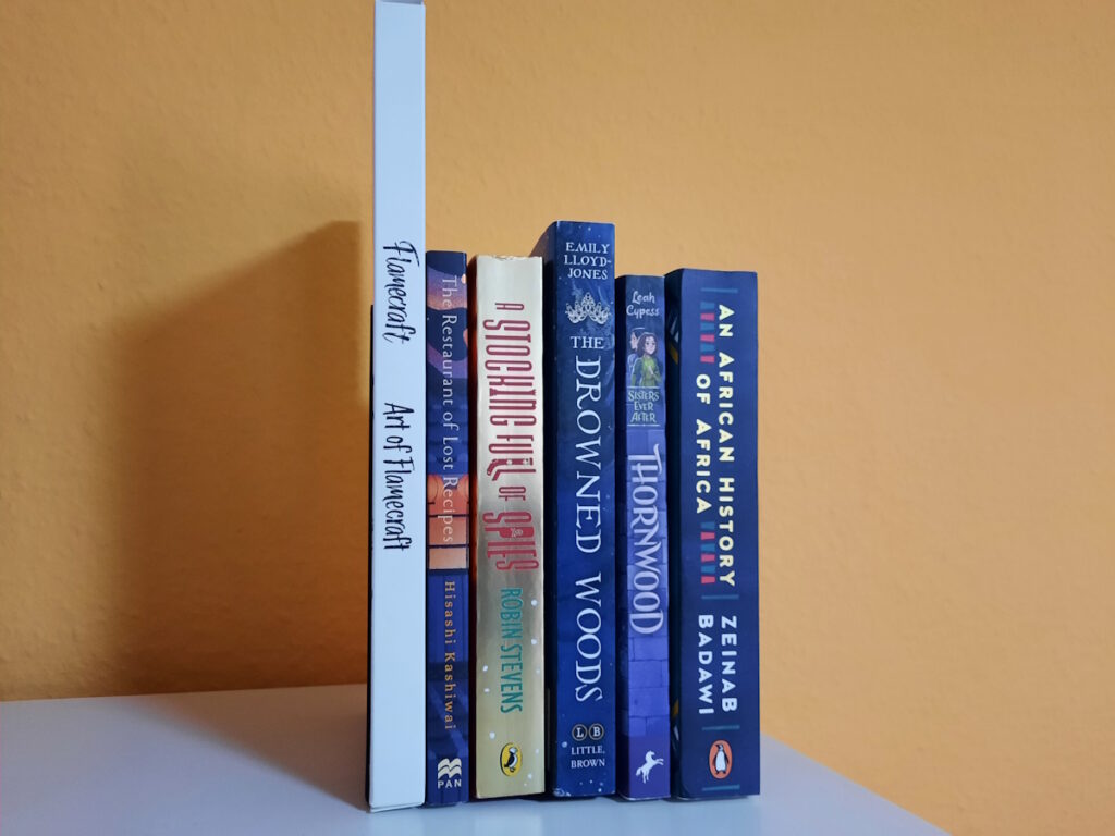 Mehrere Bücher zwischen zwei Buchstützen. Die Titel lauten von links nach rechts: Flamecraft - Art of Flamecraft von Sandara Tang, The Restaurant of Lost Recipes von Hisashi Kashiwai, A Stocking Full of Spies von Robin Stevens, The Drowned Woods von Emily Lloyd-Jones, Thornwood von Leah Cypess und An African History of Africa von Zeinab Badawi