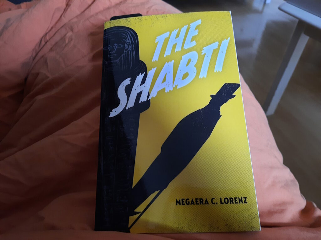 Die Taschenbuchausgabe von "The Shabti" von Megaera C. Lorenz. Das gelbe Cover zeigt auf der linken Seite den Schattenriss eines Sarkophags, dessen Schattenwurf ausschaut wie ein Mann mit Zylinder und Gehstock.