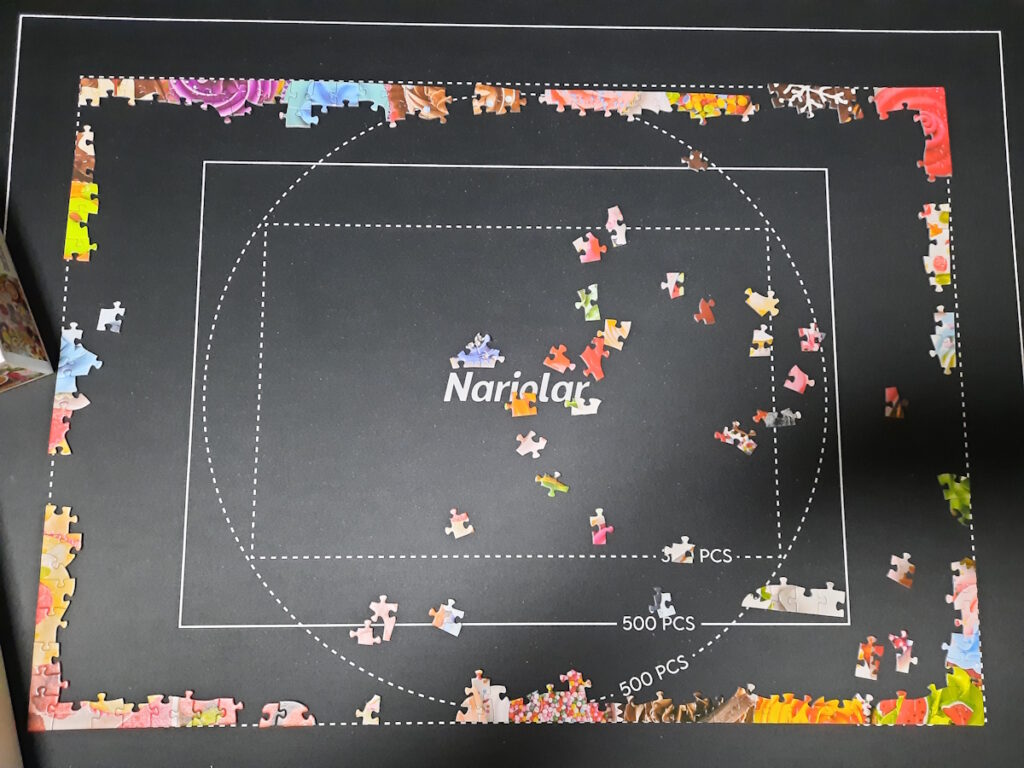 Ein angefangenes Puzzle auf einer Matte. Bislang sind gerade mal Teile des Rahmens fertig. In der Mitte der Matte liegen einige lose Teile.