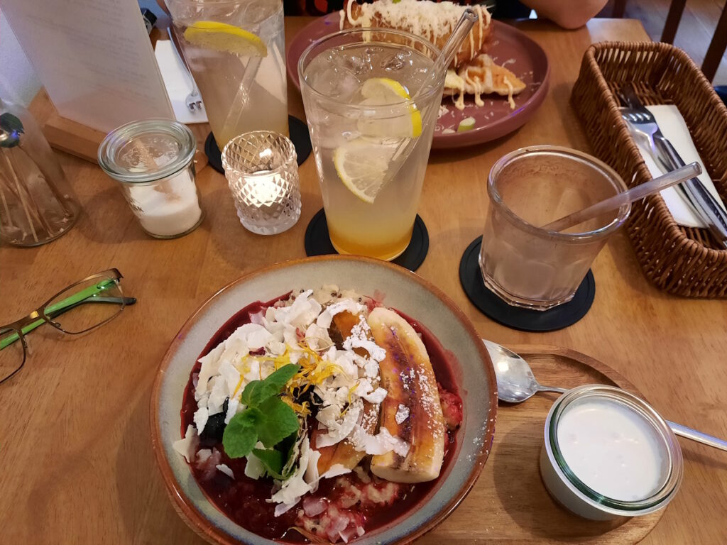 Ein Cafétisch mit mehreren Getränken. Im Vordergrund ist eine flache Schale mit einer Portion vietnamesischen Quinoa-Porridge mit karamellisierter Banane und Beerenkompott.