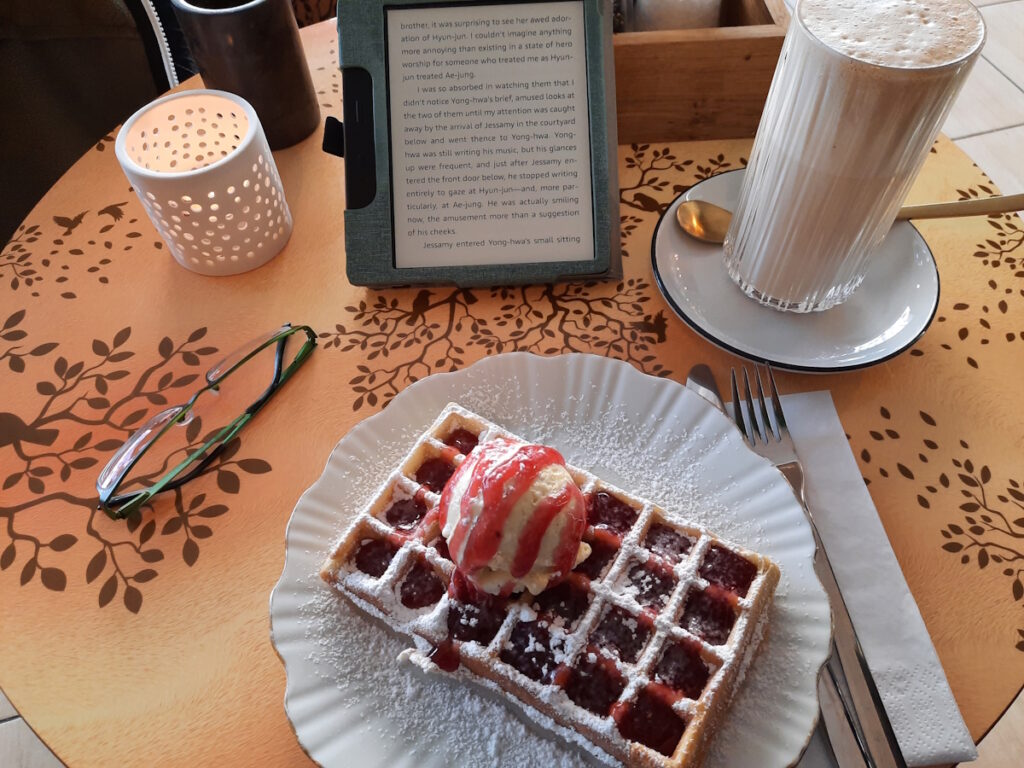Ein Cafétisch mit einer goldbraunen Waffel mit Fruchtsauce und einer Kugel Eis. Im Hintergrund sind ein Chai Latte und ein angeschalteter eReader zu erkennen.