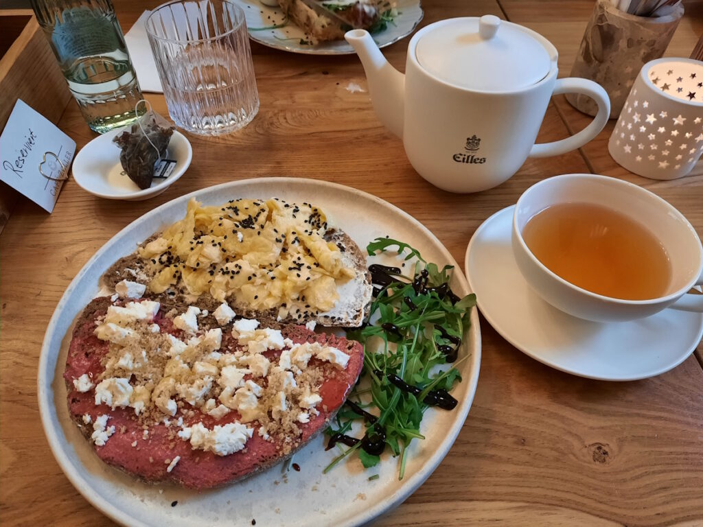 Ein Cafétisch mit einem Kännchen Tee und einem Teller mit zwei belegten Brotscheiben. Auf der einen Scheibe befindet sich ein Rote-Beete-Aufstrich, der mit Feta bestreut wurde, auf der anderen Scheibe Rührei mit Sesam bestreut.