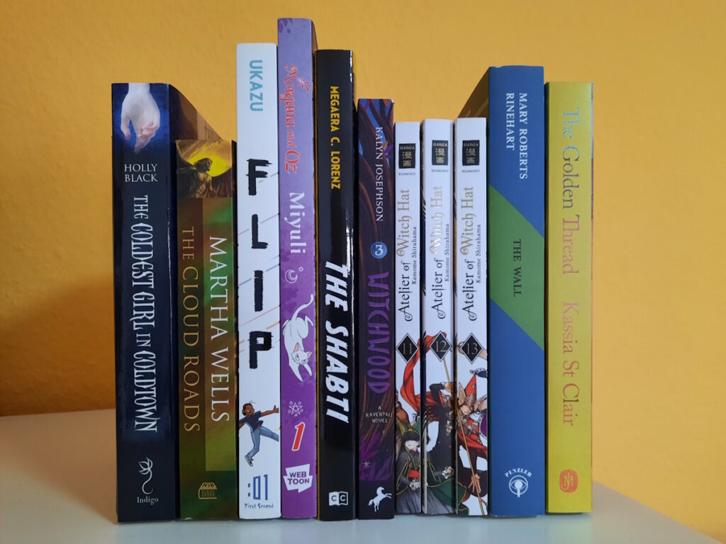 Einige Bücher, Comics und Manga zwischen zwei Bücherstützen. Die Titel lauten von links nach rechts: The Coldest Girl in Coldtown, The Cloud Road, Flip, Morgana and Oz, The Shabti, Witchwood, Atelier of Witch Hat 11, 12 und 13, The Wall und The Golden Thread.