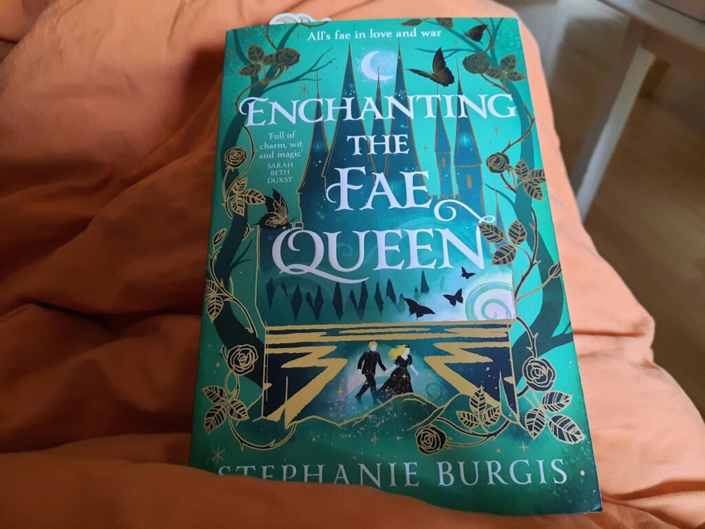 Die UK-Taschenbuchausgabe von "Enchanting the Fae Queen" von Stephanie Burgis. Das grüne Cover zeigt im unteren Drittel zwei kleine Figuren, die Hand und Hand gehen. Die eine Figur ist ein blonder Mann, die andere eine ebenfalls blonde Frau. Sie sind von goldenen Linien umgeben, die auf ein Labyrinth hindeuten. Im Hintergrund ist der Umriss eines Märchenschlosses zu sehen.
