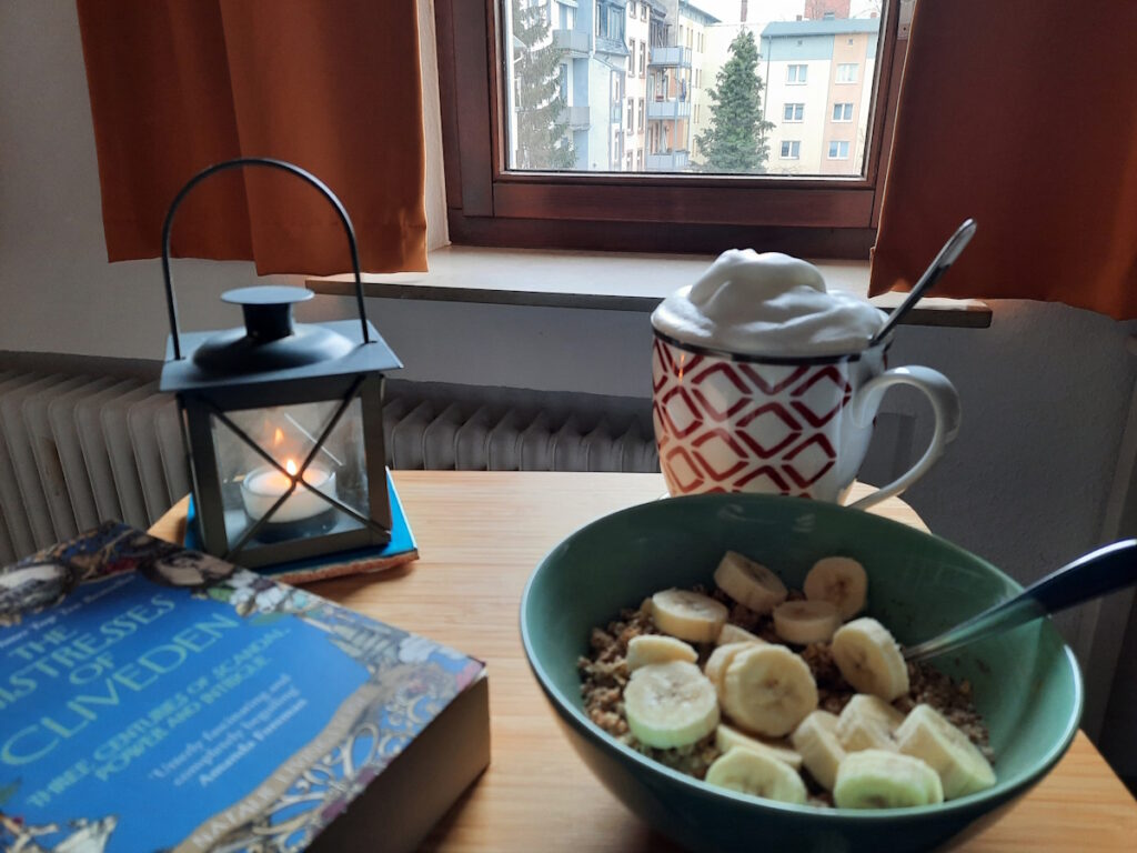 Ein kleiner Tisch mit einer zartgrünen Schale mit Porridge und Bananenscheiben. Dahinter eine große Tasse mit Milchschaumhaube. Links von der Schale lässt sich die Taschenbuchausgabe von "The Mistresses of Cliveden" von Natalie Livingstone erahnen und dahinter eine kleine brennende Sturmlaterne.