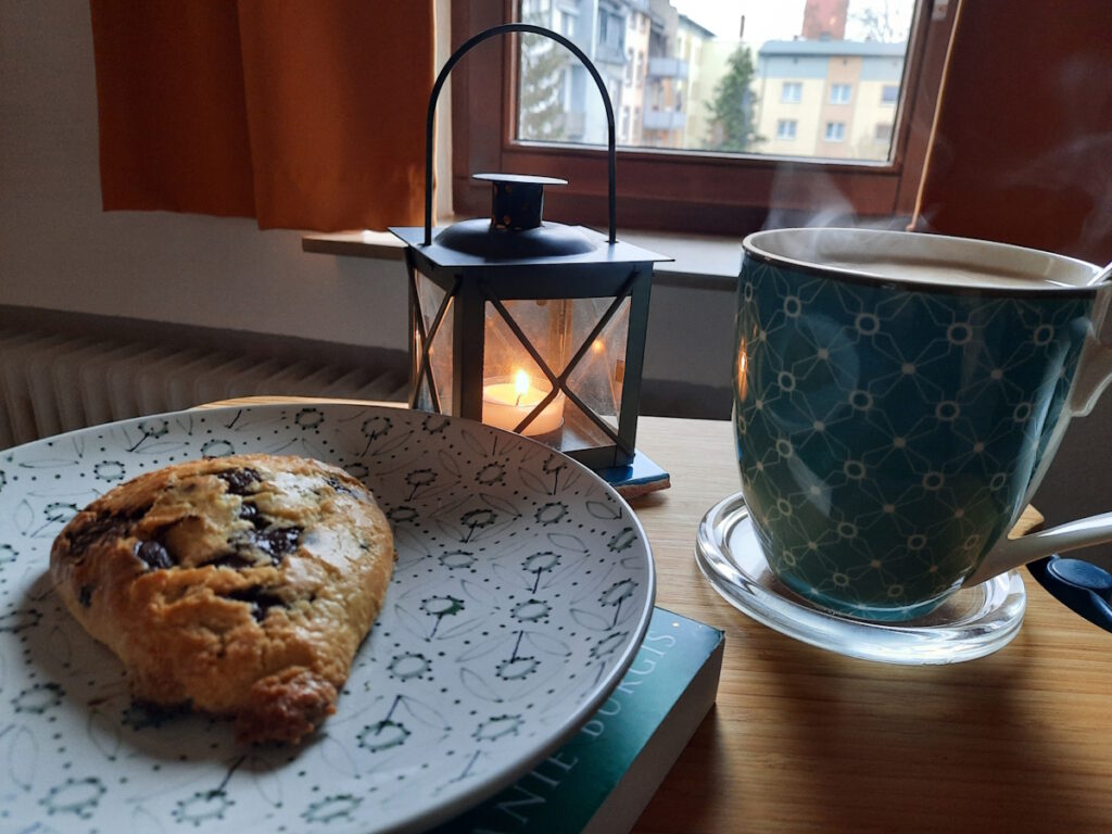 Im Vordergrund liegt ein appetitlich ausschauender goldbraun gebackener Chocolate Chip Scone auf einem gemusterten Teller. Im Hintergrund sind eine große dampfende Tasse und eine kleine brennende Sturmlaterne zu sehen.