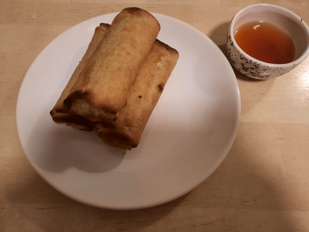 Drei Frühlingsrollen auf einem weißen Teller. Daneben eine kleine Schale mit Sweet-Chili-Sauce.