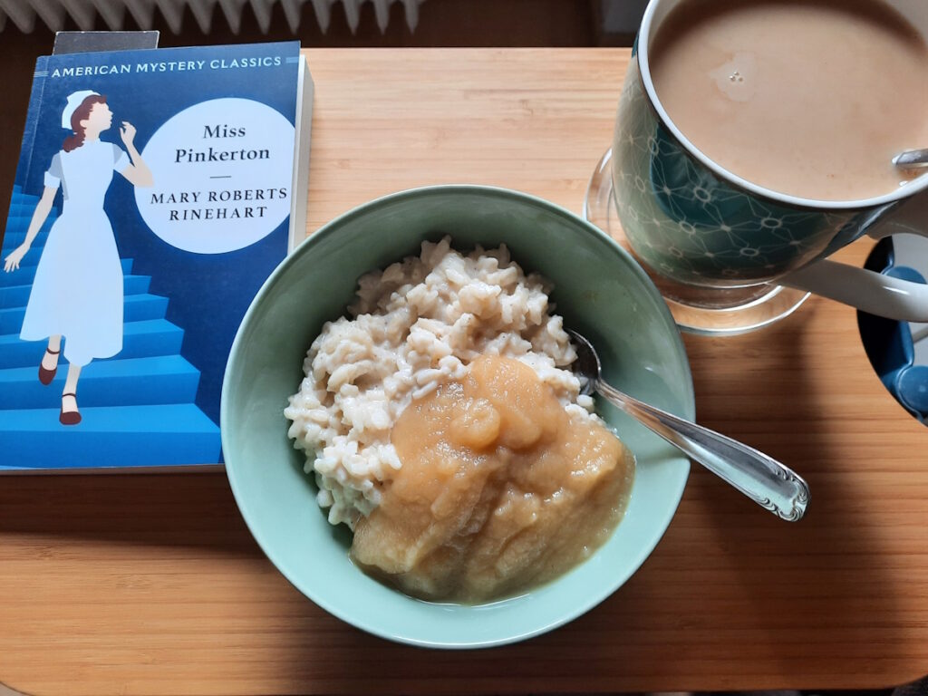 Ein kleines Tischchen mit einer Schale voller Milchreis und Apfelmus, einer großen Tasse mit Tee und Milch und der Taschenbuchausgabe von Miss Pinkerton.