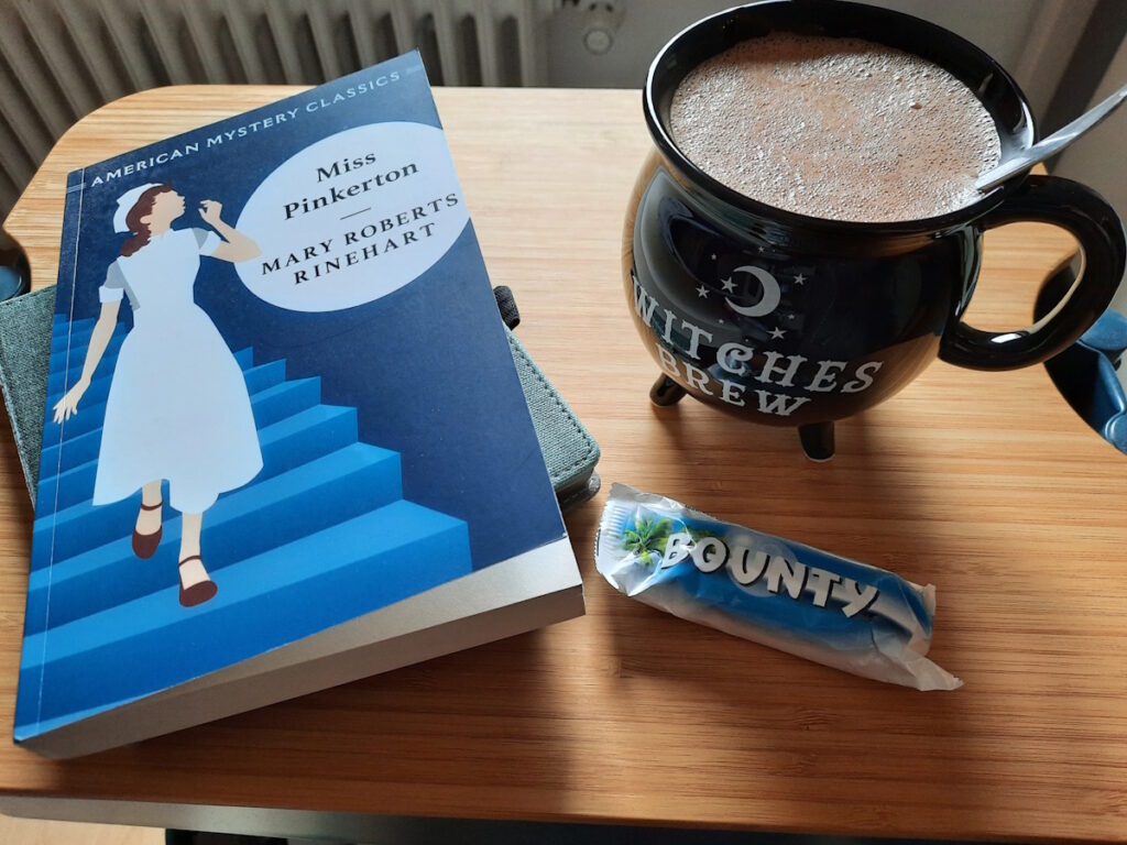 Eine Heiße Schokolade in einer schwarzen Tasse und daneben kleiner Stapel aus einem eReader und der Taschenbuchausgabe von Miss Pinkerton.