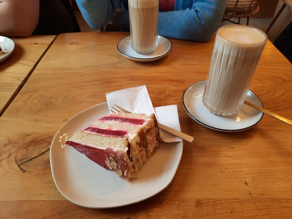 Ein Teller mit einem Stück Erdbeer-Sahne-Torte auf einem Cafétisch. Neben dem Teller steht ein Glas mit Chai Latte, im Hintergrund ist ein weiteres Glas und die dazugehörige Person zu erahnen.