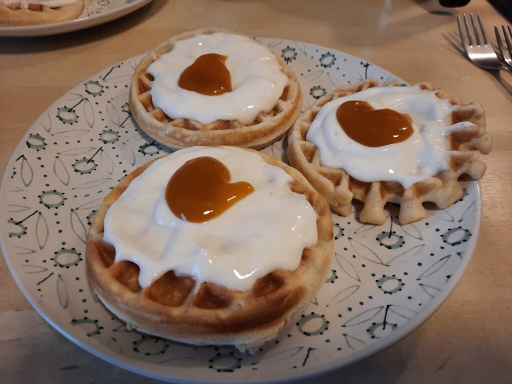 Drei kleine runde Waffeln auf einem Teller. Jede der Waffeln ist mit Vanillequark überzogen. Auf dem Quark wurde je ein Löffel Bitterorangen-Mango-Gelee gekleckst.