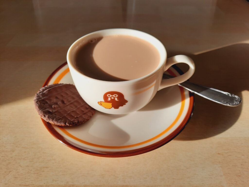 Eine Kaffeetasse mit Milchkaffee und einem Schokoladenkeks auf dem Unterteller. Das Design der Tasse ist das Gleiche wie die Tassen in Kofis Café in dem Spiel Animal Crossing. Auf der Tasse ist eine stilisierte Taube in braun mit einer gelben Tasse zu sehen. Die Untertasse hat am Rand einen braunen und einen gelben Streifen.