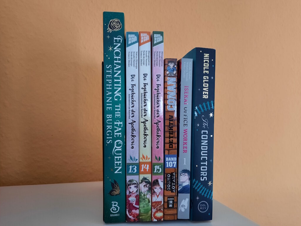 Einige Bücher und Manga zwischen zwei Buchstützen. Die Titel lauten von links nach rechts: Enchanting the Fae Queen von Stephanie Burgis, Die Tagebücher der Apothekerin 13, 14 und 15, Detektiv Conan 107, Isekai Office Worker 1 und The Conductors von Nicole Glover