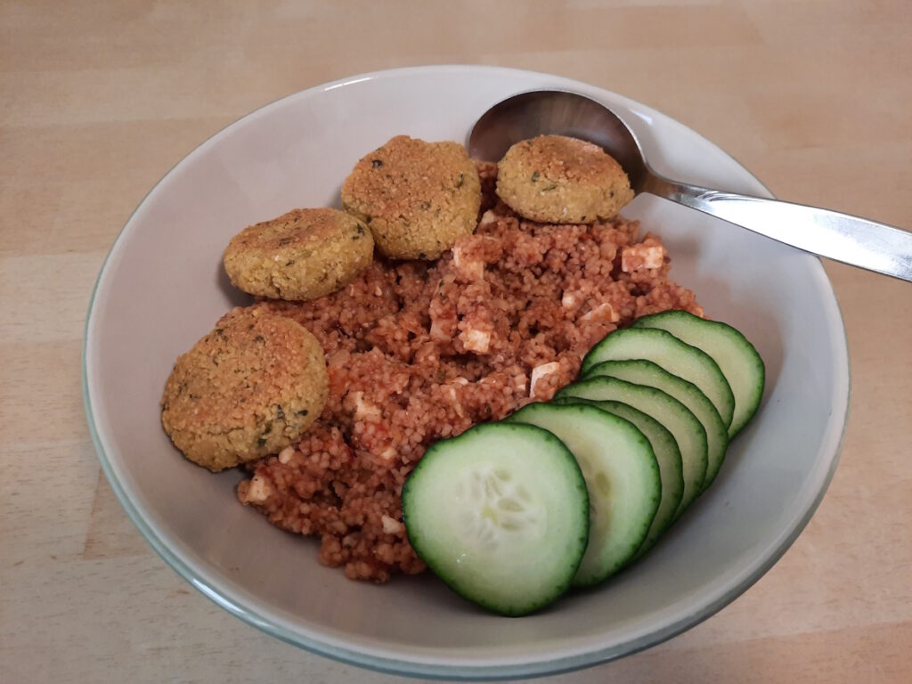 Eine Schale mit rötlichem Linsen-Couscous, vier goldbraunen Falafel und einigen Gurkenscheiben.