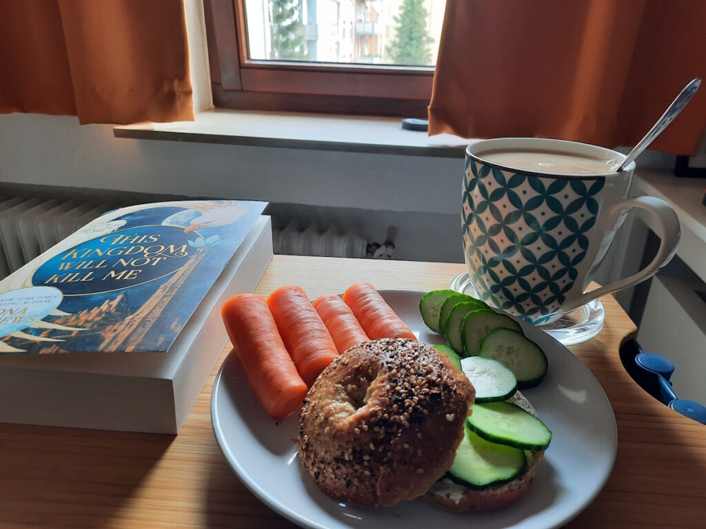Ein kleiner Tisch mit einem Teller, auf dem ein Sauerteig-Bagel mit Gurkenscheiben belegt und einige Möhrenstücke liegen. Dahinter ist eine große Tasse mit Tee zu sehen und daneben liegt die Taschenbuchausgabe von "This Kingdom Will Not Kill Me".