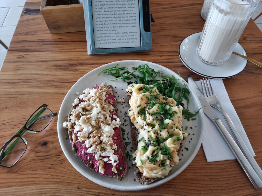 Ein Cafétisch mit zwei Scheiben Brot auf einem Teller. Die linke Scheibe wurde mit Rote-Beete-Creme, Rote Beete und Walnüssen belegt. Die rechte Scheibe mit Rührei und Kräutern. Neben dem Teller sind noch ein Glas mit Chi Latte, eine Brille, Besteck und ein angeschalteter eReader auf dem Tisch zu sehen.