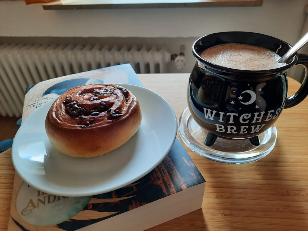 Eine Untertasse mit einer Schoko-Marzipan-Schnecke auf einem Buch. Daneben eine hexenkesselförmige Tasse mit Heißer Schokolade.
