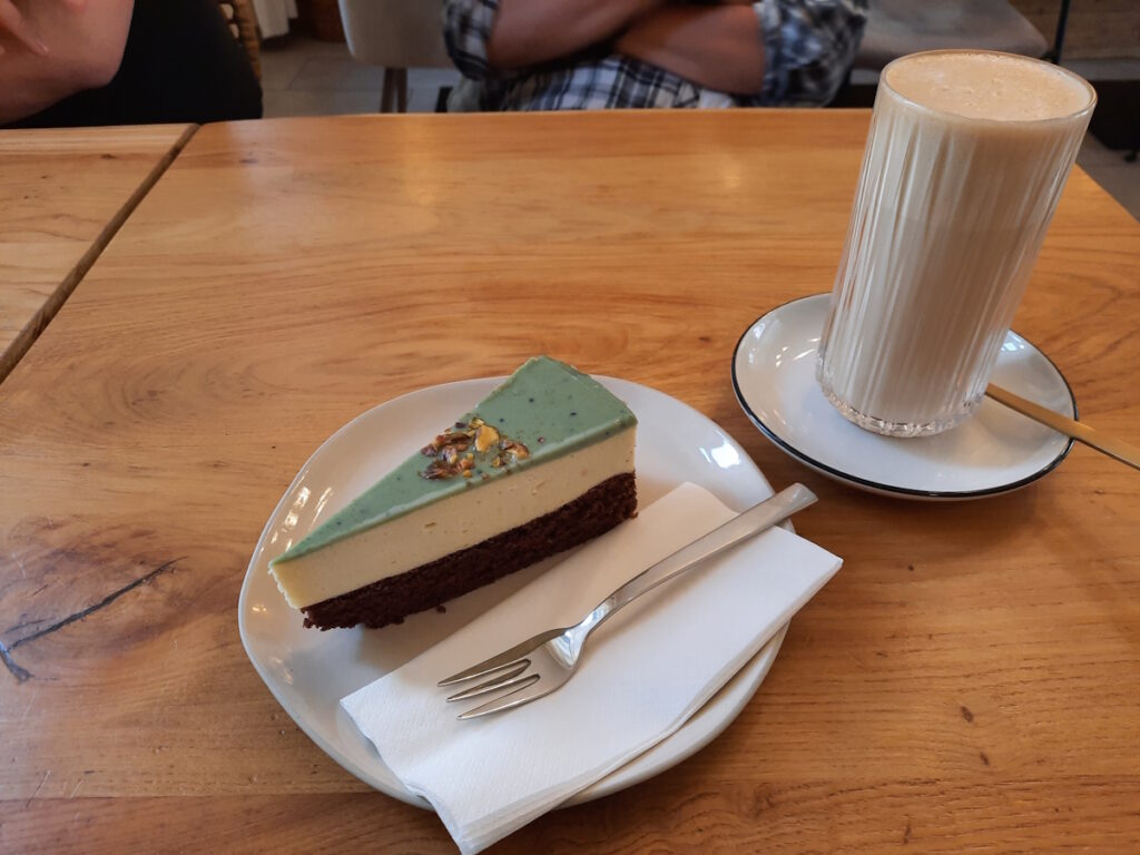 Ein Stück Parzipancreme-Torte auf einem Cafétisch. Daneben ein Glas mit einem Chai Latte.