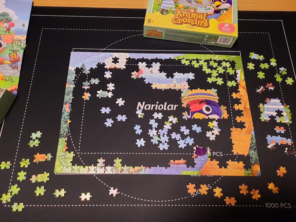 Ein angefangenes Puzzle auf einer Matte. Der Rahmen ist fertig und man kann den Kopf einer Figur erkennen, die aussieht wie ein blau-roter Vogel mit einem Strohhut. Über dem Puzzle ist ein Karton mit dem Animal-Crossing-Logo zu sehen.