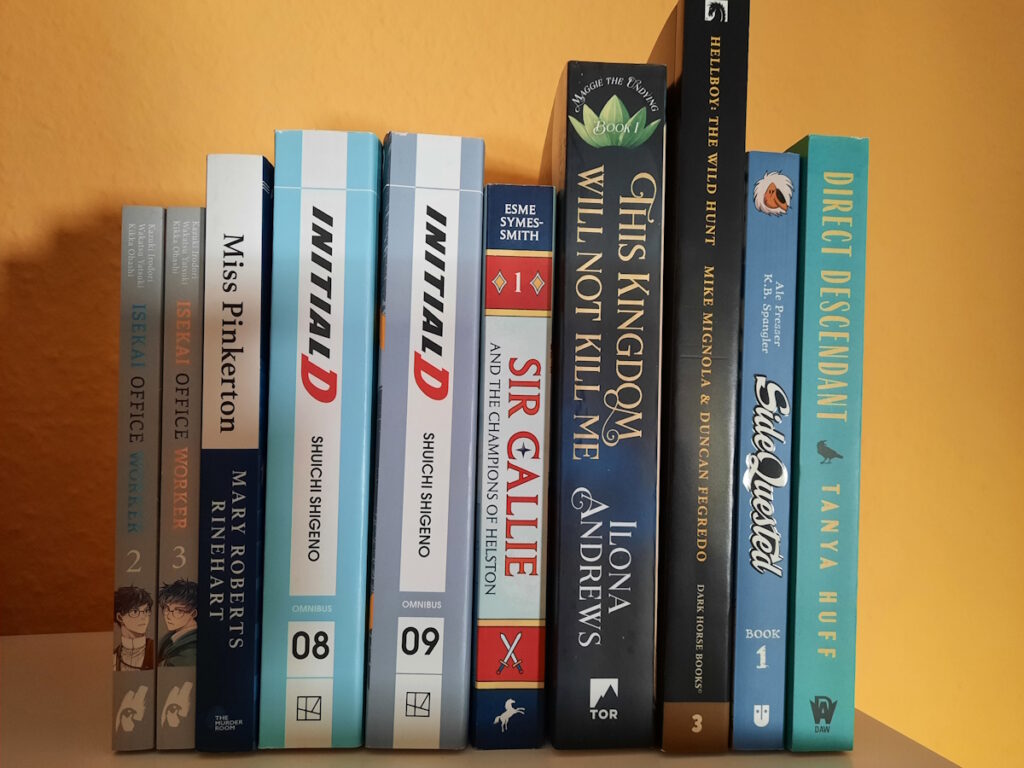 Einige Romane, Comics und Manga zwischen zwei Buchstützen. Die Titel lauten von links nach rechts: Isekai Office Worker 1+2, Miss Pinkerton, Initial D Omnibus 8+9, Sir Callie and the Champions of Helston, This Kingdom Will Not Kill Me, Hellboy: The Wild Hunt, SideQuested und Direct Descendant