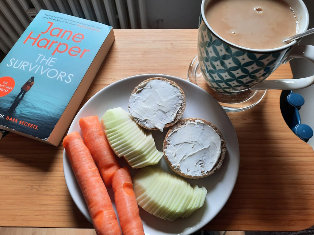 Ein Teller mit einem English Muffin (mit Frischkäse), einer in feine Scheiben geschnittenen Kohlrabi und ein paar Möhrenstückchen. Daneben eine große Tasse mit Tee und die Taschenbuchausgabe von "The Survivors" von Jane Harper.