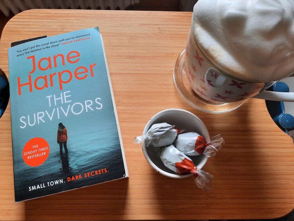Ein kleiner Tisch mit der Taschenbuchausgabe von "The Survivors", einer großen Tasse mit Milchkaffee und einer kleinen Schale mit drei Blätterkrokant-Eiern.