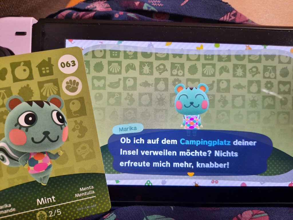 Ein Switch-Bildschirm mit der Figur Marika (ein grünes Eichhörnchen). Marika sagt: Ob ich auf dem Campingplatz deiner Insel verweilen möchte? Nichts erfreut mich mehr, knabber. Auf dem Bildschirm liegt eine Marika-Amiibo-Karte.