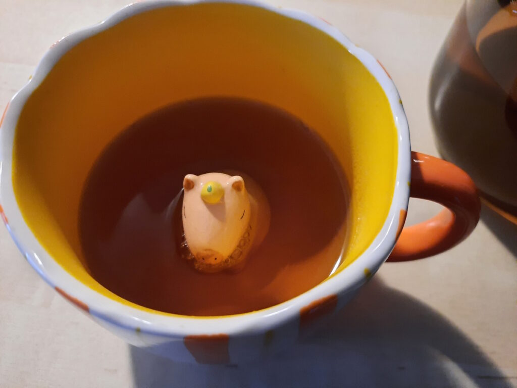 Die orangefarbende Innenansicht einer Tasse, in der ein tönernes Capybara ein Bad im Pfefferminztee nimmt.