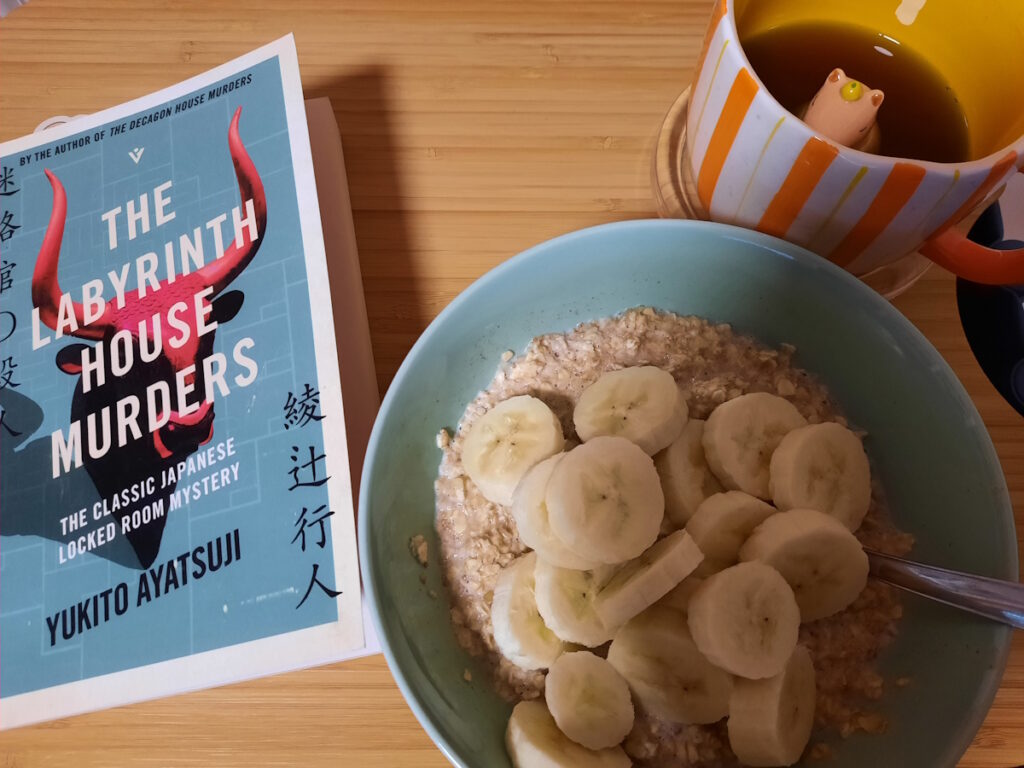 Links die zartgrüne Taschenbuchausgabe von "The Labyrinth House Murders", dessen Cover einen roten Stierkopf zeigt. Rechts eine ebenfalls zartgrüne Schale mit Porridge und Bananenscheiben.