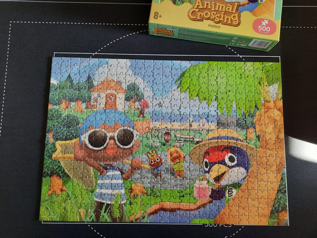 Ein kleines Puzzle mit Animal-Crossing-Motiv. Im Vordergrund sind ein blauhaariger Charakter und der Vogel Jan, im Hintergrund sieht man ein kleines Haus auf einer Klippe und mehrere Figuren an einem Strand.