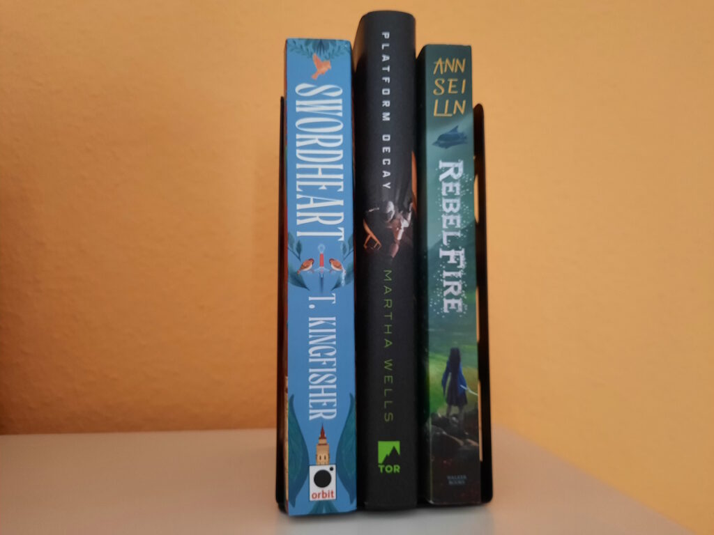 Drei Bücher zwischen zwei Buchstützen. Die Titel lauten von links nach rechts: Swordheart von T. Kingfisher, Plattform Decay von Martha Wells und Rebel Fire von Ann Sei Lin.