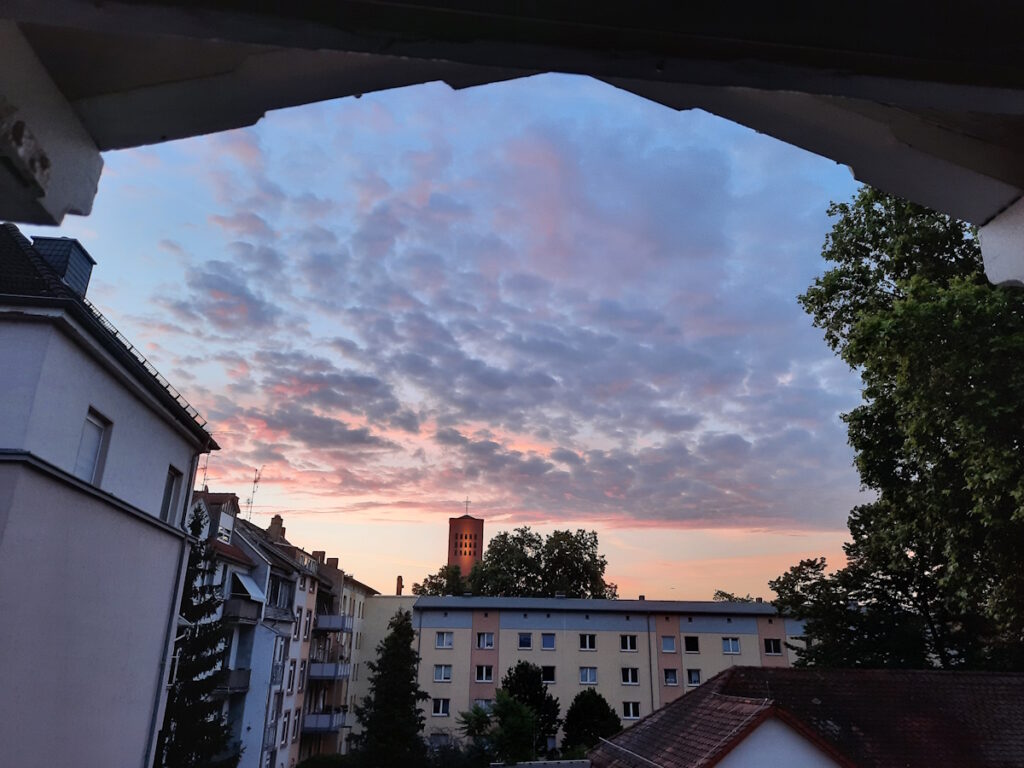 Blick aus dem Fenster auf einen Himmel, der unten rosarot und oben hellblau und mit kleinen Wolken beflockt ist. Die Sonne ist noch nicht aufgegangen, weshalb die Häuser im Hintergrund noch im Schatten liegen.