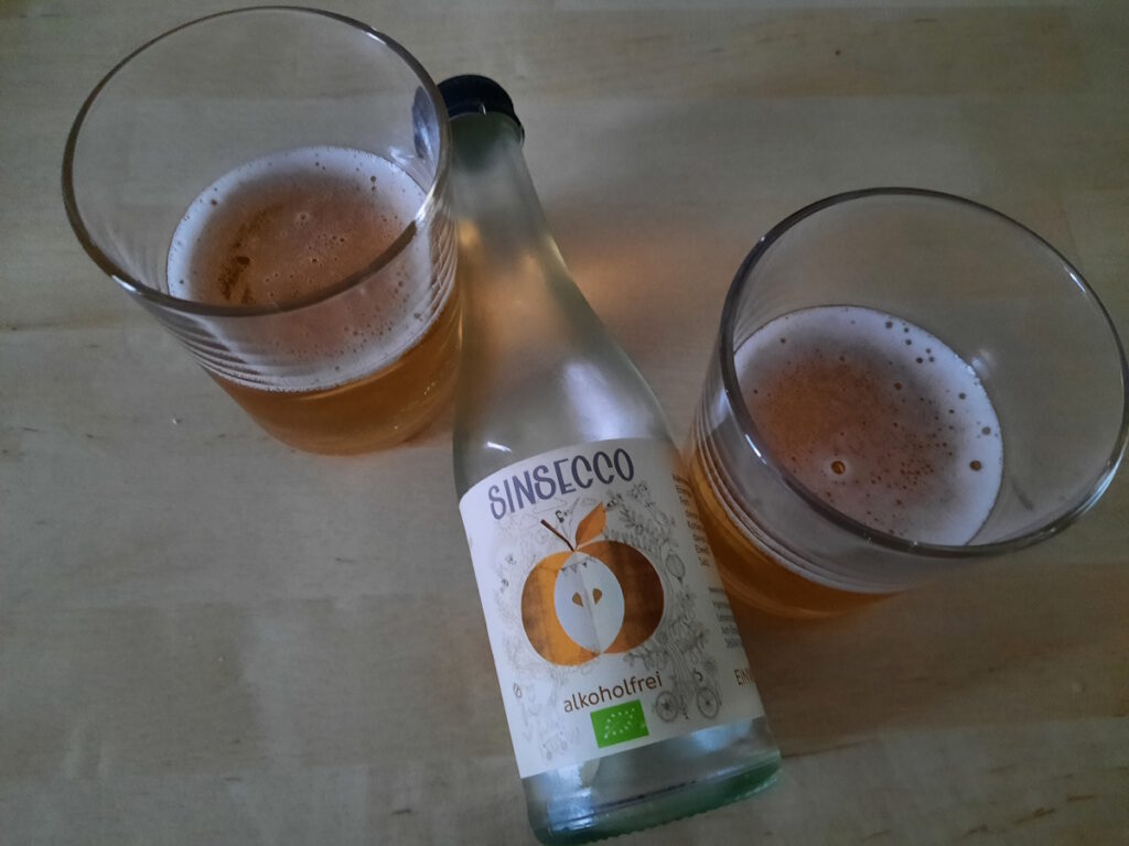Eine kleine Flasche Sinsecco zwischen zwei halb gefüllten Trinkgläsern. Die Flasche wirbt damit alkoholfrei zu sein und das Etikett zeigt einen angeschnittenen Apfel.